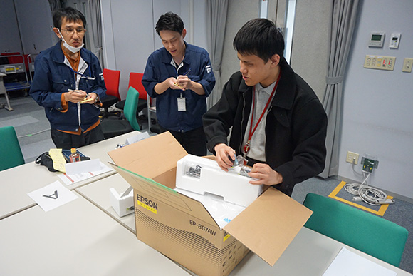 プリンターの個装箱を開けている人と観察する人たち